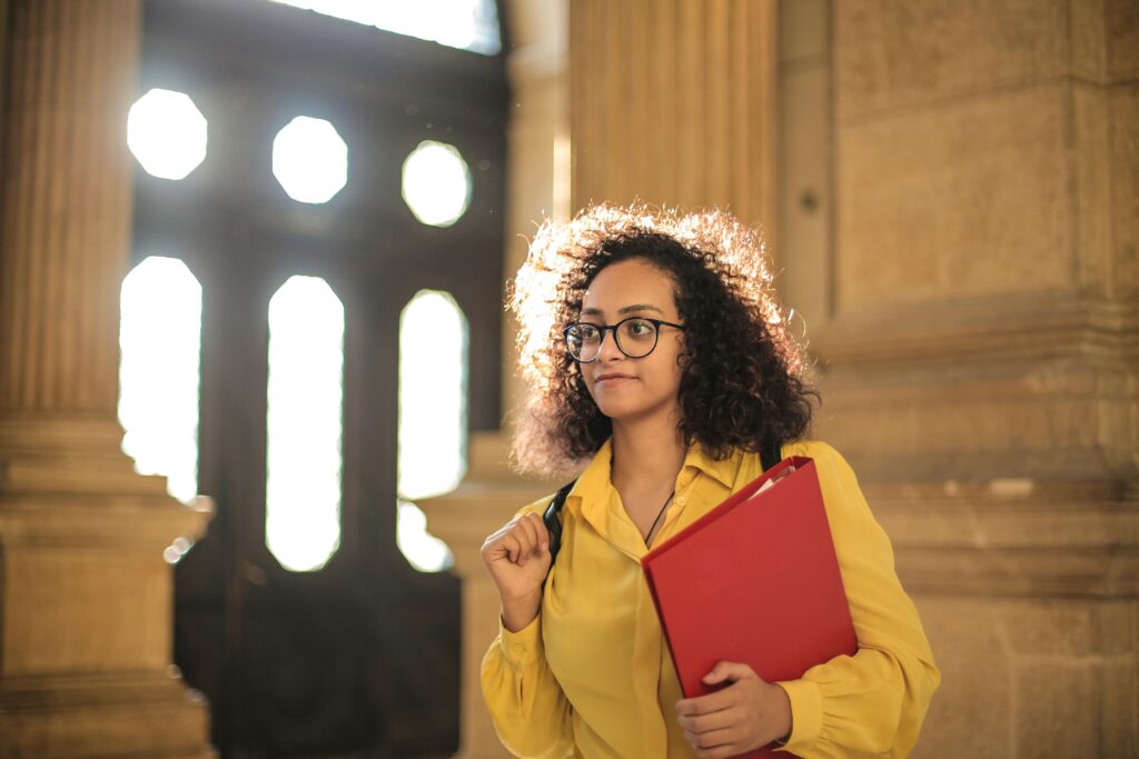 pexels-photo-3762803-3762803 A young woman in a library carrying a red binder, bathed in sunlight.
